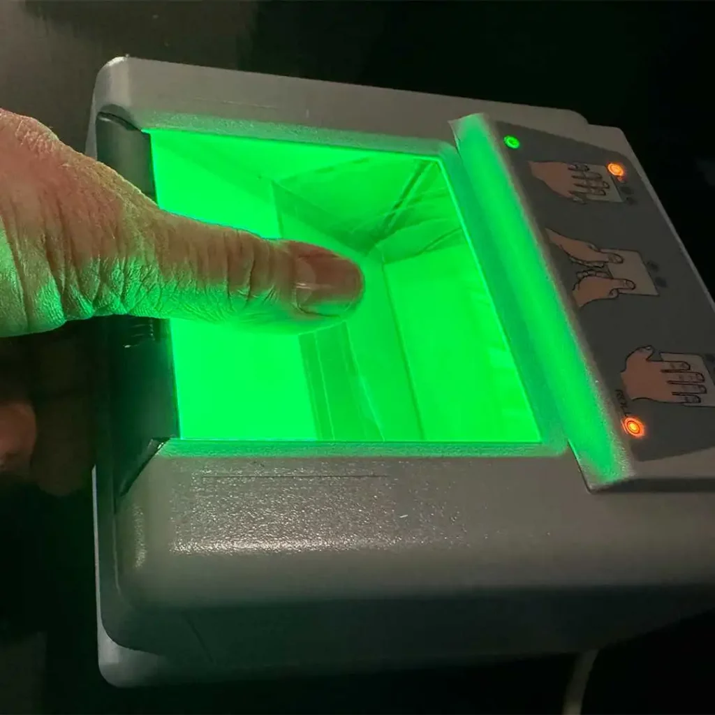 A person's hand using a LiveScan machine, capturing a digital fingerprint for a background check. The device glows with a green light.